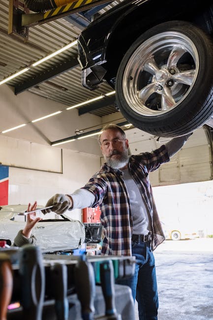 Bearded mechanic working on a vehicle in an auto repair shop, handing a tool to an assistant.